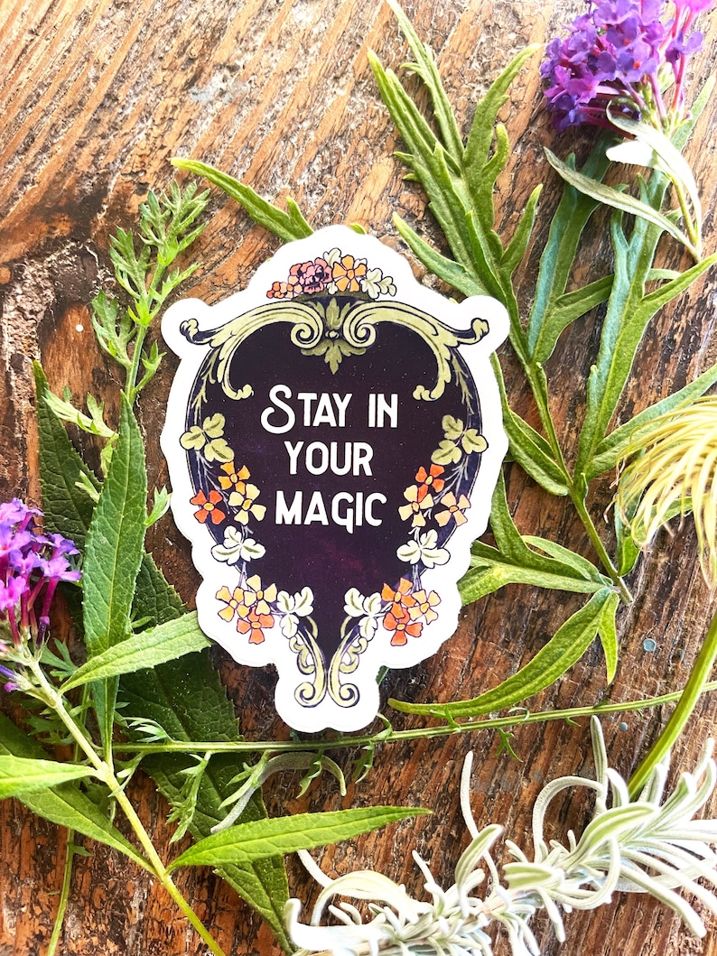 May include: A sticker with a dark purple background and gold accents. The sticker features a decorative frame with floral details and the text "Stay in your magic".