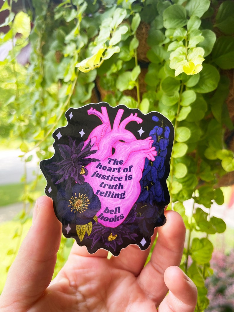 Puede incluir: Una pegatina de coraz&oacute;n anat&oacute;mico rosa con detalles florales negros y la cita "El coraz&oacute;n de la justicia es decir la verdad, bell hooks" sobre un fondo negro.