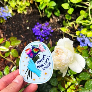 May include: A blue and white sticker with a rainbow bouquet of roses and a gloved hand holding the stems. The sticker says "Support Your Local Queers".