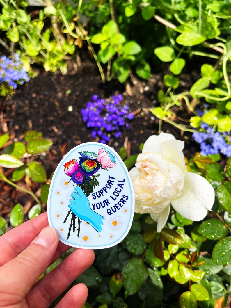 May include: A blue and white sticker with a rainbow bouquet of roses and a gloved hand holding the stems. The sticker says "Support Your Local Queers".