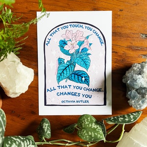 Peut inclure: Une fleur rose et blanche avec des feuilles vertes dans un cadre bleu et blanc avec le texte "ALL THAT YOU TOUCH, YOU CHANGE. ALL THAT YOU CHANGE, CHANGES YOU OCTAVIA BUTLER".