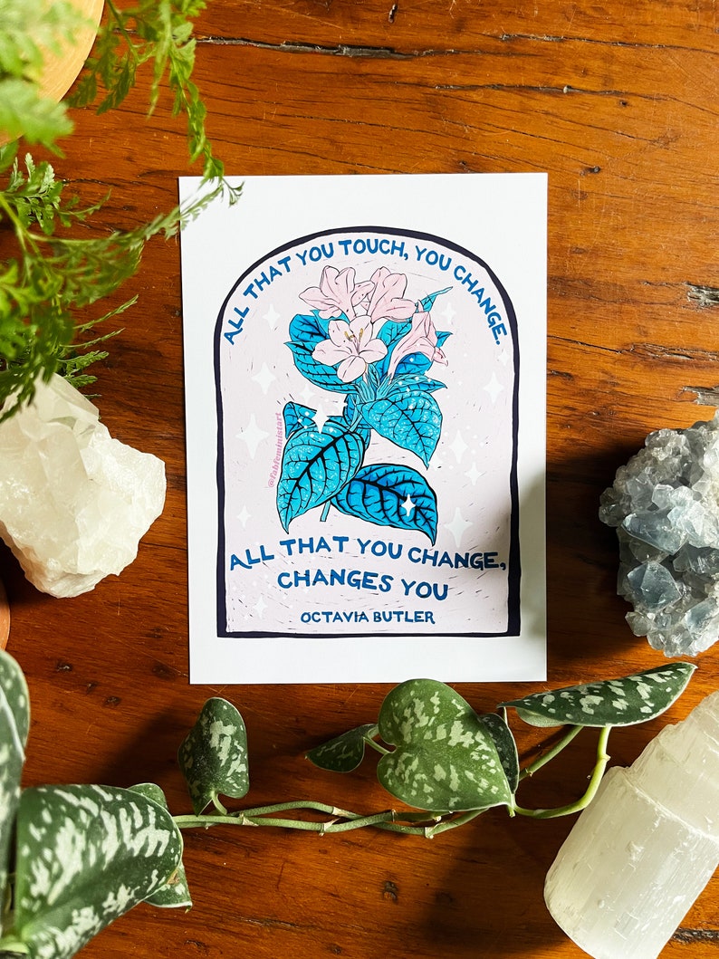 Peut inclure: Une fleur rose et blanche avec des feuilles vertes dans un cadre bleu et blanc avec le texte "ALL THAT YOU TOUCH, YOU CHANGE. ALL THAT YOU CHANGE, CHANGES YOU OCTAVIA BUTLER".