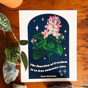 May include: A navy blue print with a white archway featuring a pink flower with green leaves and white stars. The quote "The function of freedom is to free someone else. - Toni Morrison" is printed below the flower.