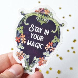 May include: A sticker with a dark blue background and a white border. The sticker features a floral design with orange and green flowers. The text "Stay in your magic" is written in white on the sticker.