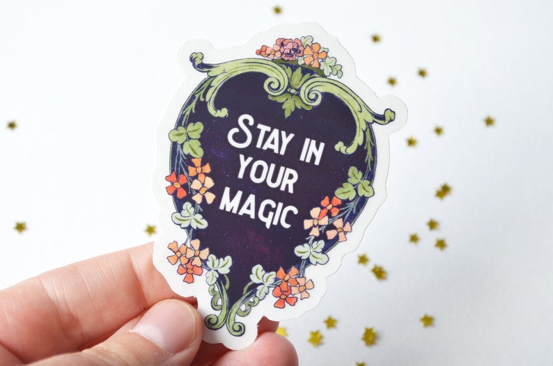 May include: A sticker with a dark blue background and a white border. The sticker features a floral design with orange and green flowers. The text "Stay in your magic" is written in white on the sticker.
