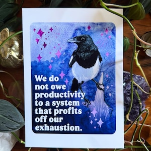 May include: A white card with a watercolor painting of a black and white magpie bird against a blue and purple background. The card has the text: "We do not owe productivity to a system that profits off our exhaustion."