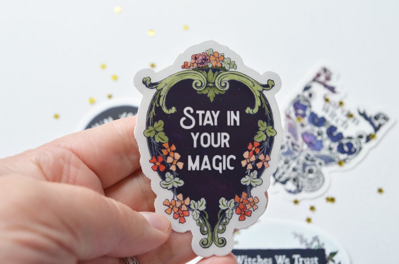 May include: A black and white sticker with a floral border and the text "Stay in your magic" in white lettering.