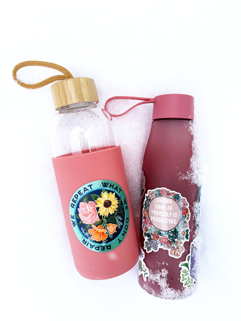 May include: Two pink water bottles with different designs. The bottle on the left has a sticker with flowers and the text "We repeat what we don't repair". The bottle on the right has a sticker with flowers and the text "Taking care of yourself is productive".