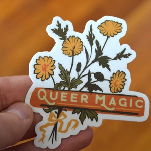 Puede incluir: Una pegatina blanca con un ramo de flores amarillas y hojas verdes. Las flores tienen centros naranjas. La pegatina tiene una pancarta con el texto "Queer Magic" en letras marrones sobre un fondo naranja.
