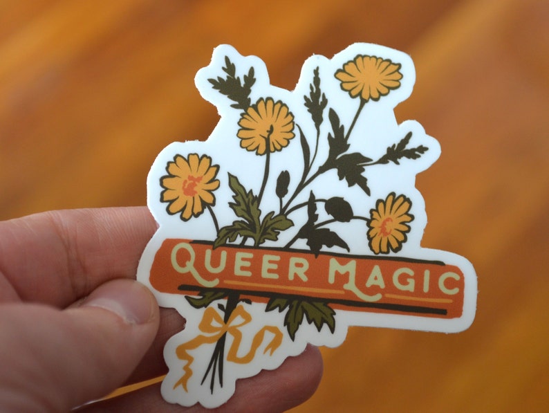 Puede incluir: Una pegatina blanca con un ramo de flores amarillas y hojas verdes. Las flores tienen centros naranjas. La pegatina tiene una pancarta con el texto "Queer Magic" en letras marrones sobre un fondo naranja.