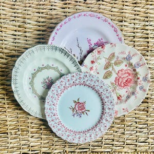 May include: A collection of five vintage ceramic plates with floral patterns. The plates feature various designs, including roses, sprigs, and decorative borders in shades of pink, green, and white. The plates are arranged on a woven wicker surface.