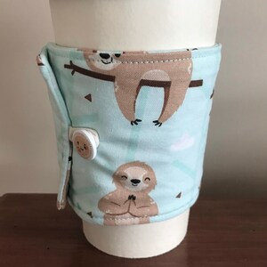 May include: A reusable coffee cup sleeve featuring a sloth pattern on a light blue background. The sleeve is secured with a button. The cup has a black lid.