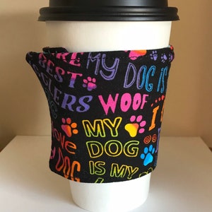May include: A white paper coffee cup with a black lid, wrapped with a black fabric sleeve. The sleeve is printed with colorful text and paw prints that read "My Dog is My Best Friend".
