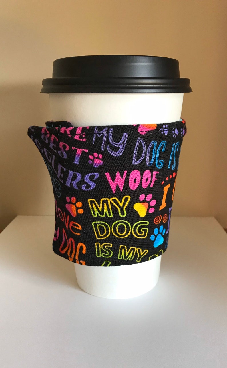 May include: A white paper coffee cup with a black lid, wrapped with a black fabric sleeve. The sleeve is printed with colorful text and paw prints that read "My Dog is My Best Friend".