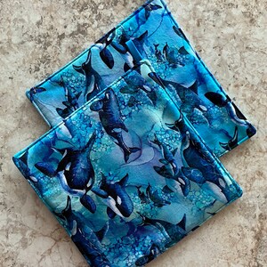 May include: Two blue and black patterned pot holders with a blue and white watercolour design of orcas swimming in a blue ocean. The pot holders have a loop for hanging.
