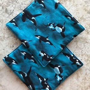 Può includere: Due presine quadrate con sfondo blu e un motivo di orche nere e bianche. Le presine hanno un anello nero per appenderle e un bordo blu.