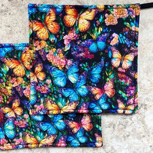 Peut inclure: Deux maniques carrées avec un motif vibrant de papillons et de fleurs. Le motif présente des papillons orange, jaunes, bleus et roses sur un fond noir avec un feuillage vert et des fleurs colorées. Chaque manique a une boucle noire.