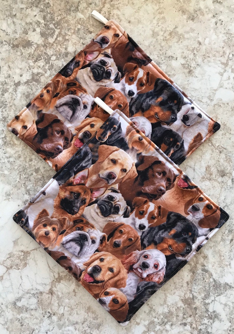 May include: Two brown and white pot holders with a pattern of various dog breeds. The pot holders are made of fabric and have a white loop for hanging.