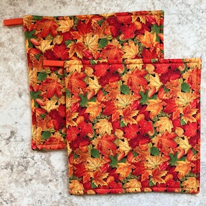 May include: Two red, orange and yellow quilted pot holders with a fall leaf pattern. The pot holders have a loop for hanging.