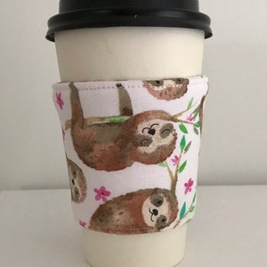 May include: A white paper coffee cup with a black lid, wrapped with a fabric sleeve. The sleeve features a repeating pattern of brown sloths hanging from tree branches with pink flowers.