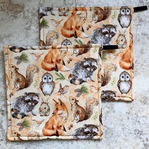 May include: Two beige kitchen pot holders with a woodland animal print. The print features foxes, raccoons, owls, squirrels, and rabbits in shades of brown, grey, and white on a beige background.