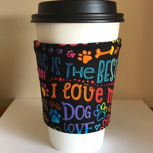 May include: A white paper coffee cup with a black lid, wrapped with a black fabric sleeve. The sleeve is printed with colorful text and paw prints that read "DOG IS THE BEST I LOVE MY DOG".