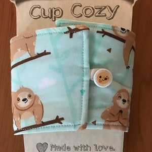 May include: A fabric cup cosy with a light blue background and a pattern of brown sloths on branches. The cosy has a button closure with a smiling sun design. The words "Cup Cozy" are printed on the top.