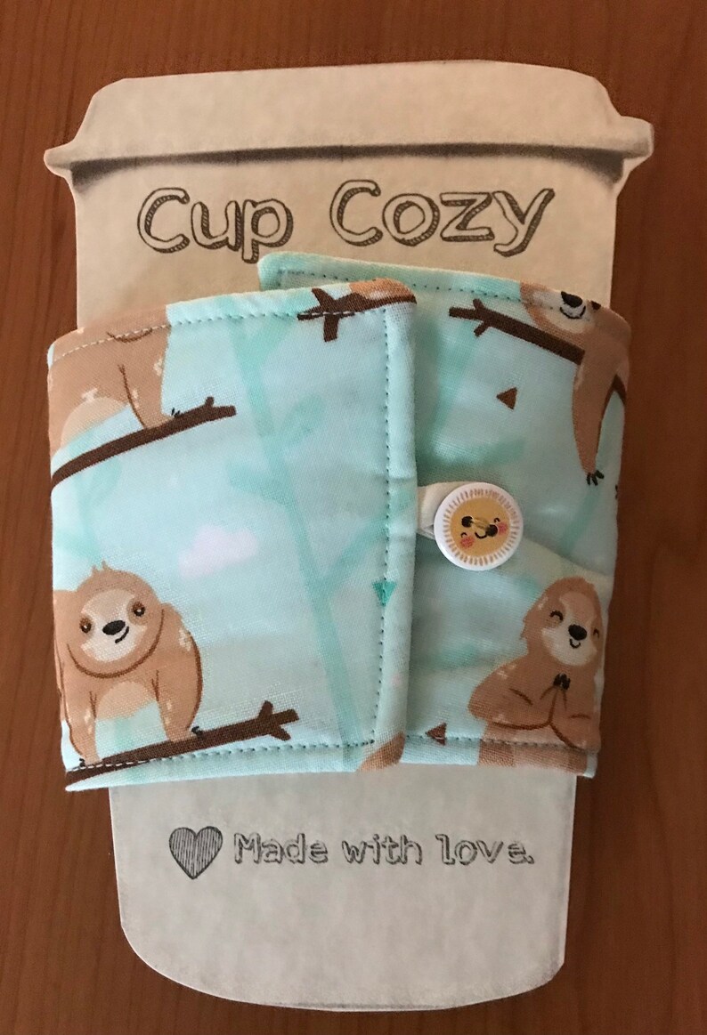 May include: A fabric cup cosy with a light blue background and a pattern of brown sloths on branches. The cosy has a button closure with a smiling sun design. The words "Cup Cozy" are printed on the top.