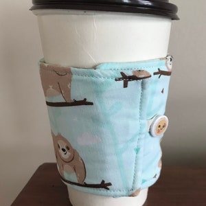 May include: A white paper coffee cup with a black lid, wrapped in a light blue fabric sleeve. The sleeve features a repeating pattern of brown sloths on branches. A button with a smiling sun secures the sleeve.