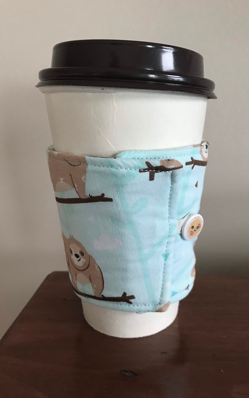 May include: A white paper coffee cup with a black lid, wrapped in a light blue fabric sleeve. The sleeve features a repeating pattern of brown sloths on branches. A button with a smiling sun secures the sleeve.