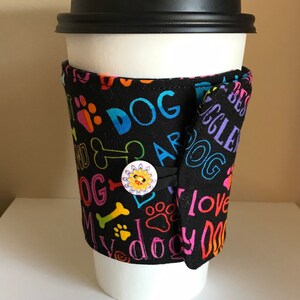 May include: A white paper coffee cup with a black lid, wrapped in a black fabric sleeve. The sleeve is decorated with colorful paw prints, bones, and the words "DOG" and "I love my dog". A button with a sun design secures the sleeve.