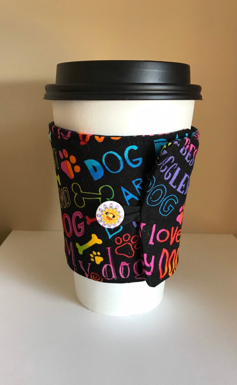 May include: A white paper coffee cup with a black lid, wrapped in a black fabric sleeve. The sleeve is decorated with colorful paw prints, bones, and the words "DOG" and "I love my dog". A button with a sun design secures the sleeve.