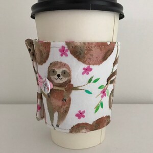 May include: A white coffee cup cozy with a sloth design. The cozy has a pink button closure and features sloths hanging from branches with pink flowers.