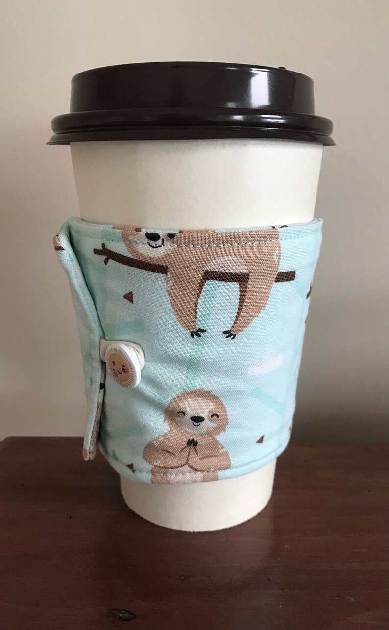 May include: A reusable coffee cup sleeve featuring a sloth pattern on a light blue background. The sleeve is secured with a button. The cup has a black lid.