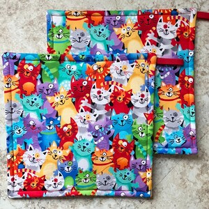 Peut inclure: Deux maniques à motifs de chats colorés. Les maniques présentent un motif répété de chats de dessins animés dans diverses couleurs, notamment le rouge, l'orange, le bleu, le vert et le violet. Chaque manique a une boucle rouge pour l'accrocher.