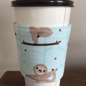 May include: A white paper coffee cup with a black lid, encased in a light blue fabric sleeve. The sleeve displays a repeating pattern of brown sloths on branches. The sloth design adds a charming, whimsical element.