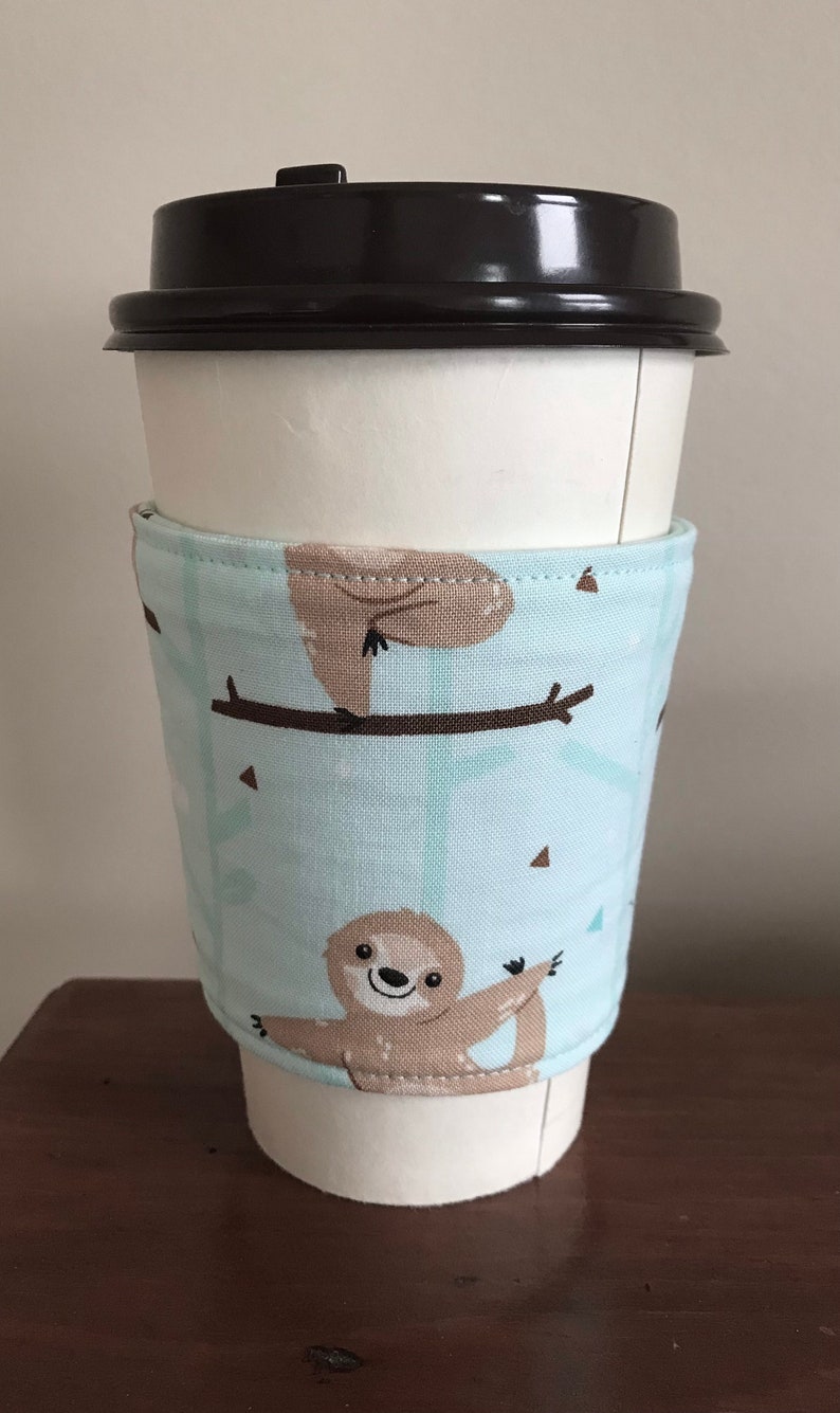 May include: A white paper coffee cup with a black lid, encased in a light blue fabric sleeve. The sleeve displays a repeating pattern of brown sloths on branches. The sloth design adds a charming, whimsical element.