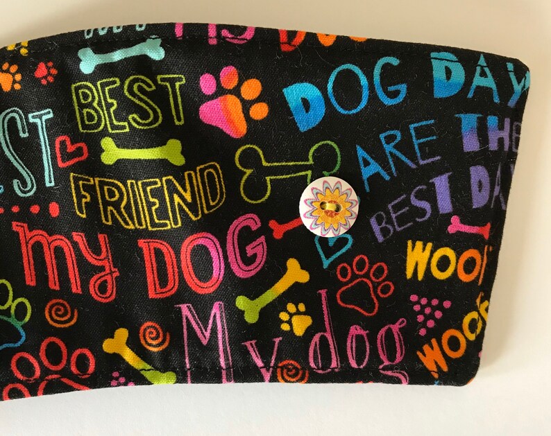 May include: Black fabric coffee cup sleeve with colorful text and dog-themed graphics. The text includes "Best Friend," "My Dog," and "Dog Day." A button with a floral design secures the sleeve.