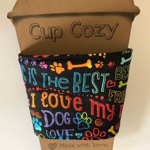 May include: A brown cardboard cup cozy with the words "Cup Cozy" printed on it. The black fabric wrap features colorful paw prints, bones, and the text "I love my dog". The bottom of the cozy says "Made with love".