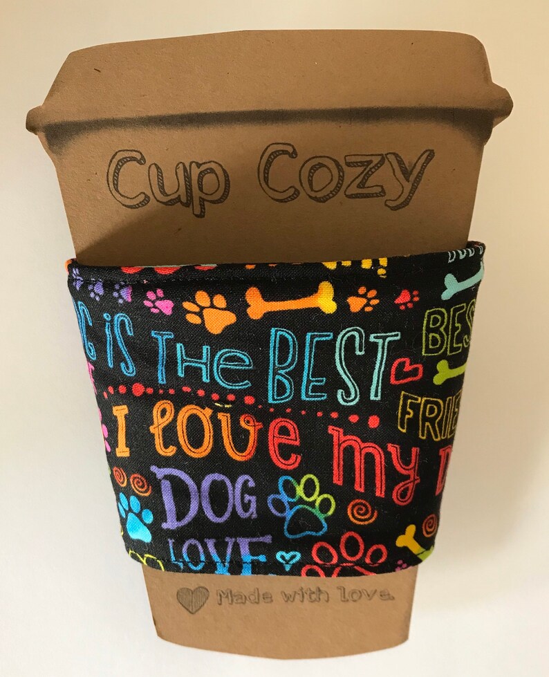 May include: A brown cardboard cup cozy with the words "Cup Cozy" printed on it. The black fabric wrap features colorful paw prints, bones, and the text "I love my dog". The bottom of the cozy says "Made with love".