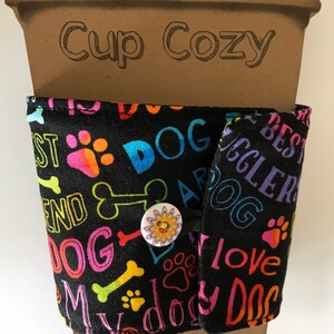 May include: Black fabric cup cozy with colorful dog-themed text and paw prints. The text includes "DOG", "BEST", "FRIEND", and "LOVE". A button closure with a floral design secures the cozy around a brown paper cup. The words "Cup Cozy" are printed on the cup.