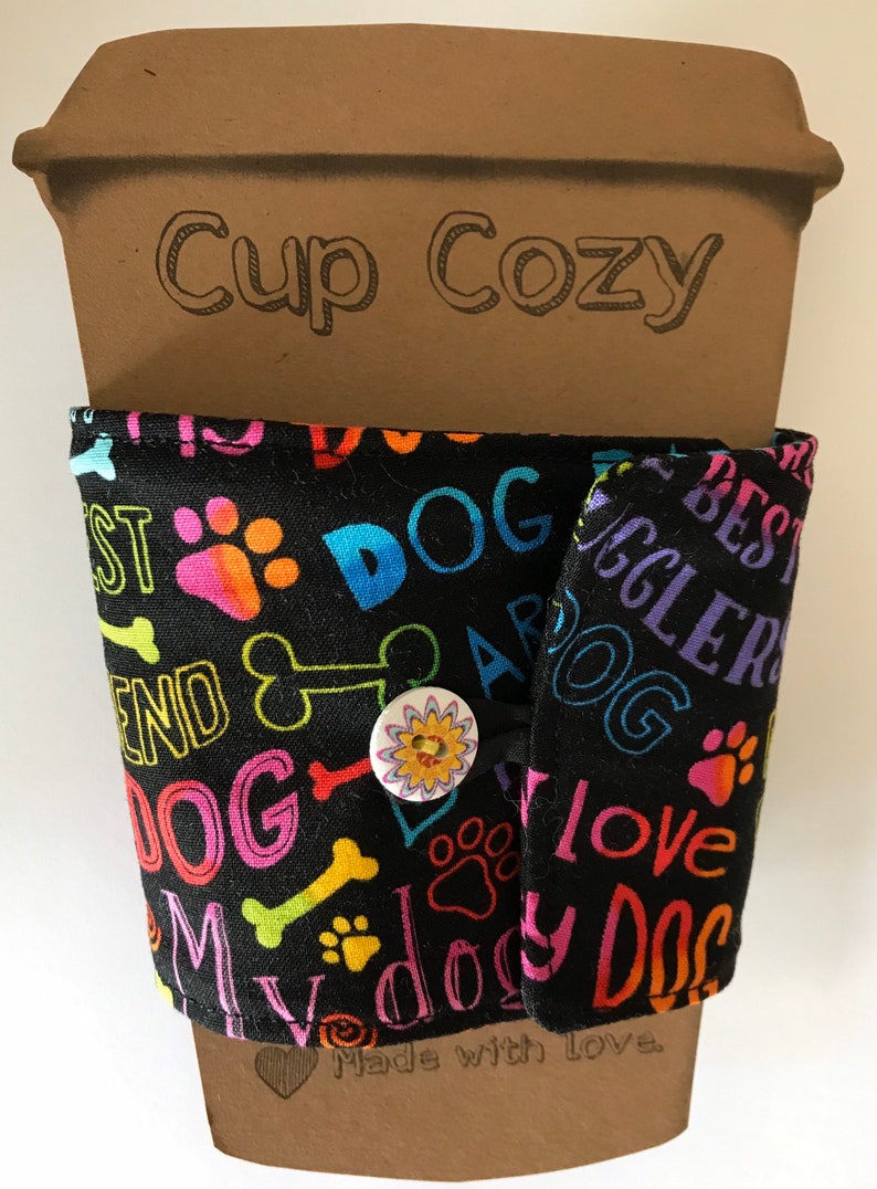 May include: Black fabric cup cozy with colorful dog-themed text and paw prints. The text includes "DOG", "BEST", "FRIEND", and "LOVE". A button closure with a floral design secures the cozy around a brown paper cup. The words "Cup Cozy" are printed on the cup.