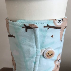 May include: A white paper coffee cup with a black lid, encased in a light blue fabric sleeve. The sleeve has a pattern of brown sloths and fastens with a button. The sleeve is designed to keep the coffee warm.