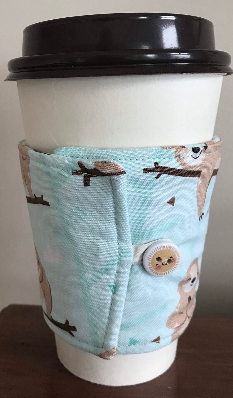 May include: A white paper coffee cup with a black lid, encased in a light blue fabric sleeve. The sleeve has a pattern of brown sloths and fastens with a button. The sleeve is designed to keep the coffee warm.