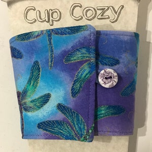 May include: A fabric cup cozy with a blue and purple dragonfly pattern. The cozy is secured with a button and is attached to a cardboard cutout that reads "Cup Cozy" and "Made with love."