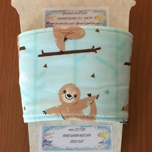 May include: A light blue coffee cup sleeve featuring a repeating pattern of brown sloths on branches. The sleeve is made of 100% cotton and has the text "HAND WASH AND DRY IRON FLAT".