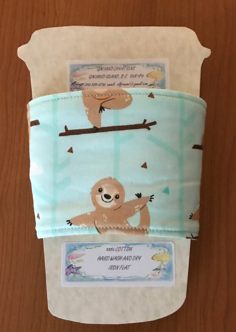 May include: A light blue coffee cup sleeve featuring a repeating pattern of brown sloths on branches. The sleeve is made of 100% cotton and has the text "HAND WASH AND DRY IRON FLAT".