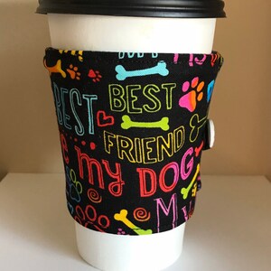 May include: A black coffee cup sleeve with the words "BEST FRIEND" and "I love my DOG" in colorful lettering. The sleeve features paw prints and bone designs. A white button secures the sleeve.