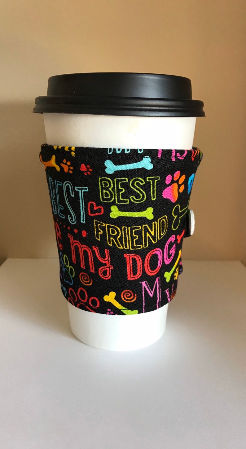 May include: A black coffee cup sleeve with the words "BEST FRIEND" and "I love my DOG" in colorful lettering. The sleeve features paw prints and bone designs. A white button secures the sleeve.