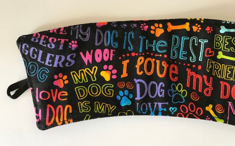 May include: Black fabric with colorful text and paw prints. The text includes phrases like "My Dog is the Best," "I Love My Dog," and "Woof." A black loop is attached to the side.